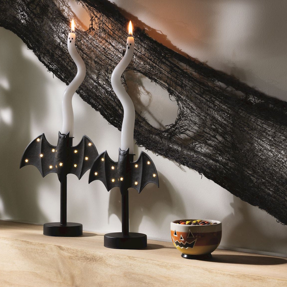Two ghost candles in bat-shaped candlesticks on a shelf with Halloween decor.