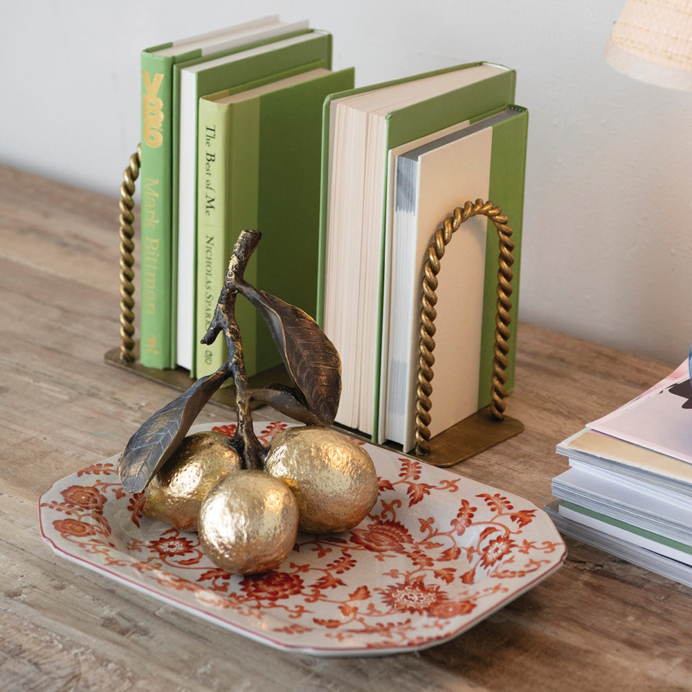 Gold twisted-metal arched bookends holding green books on a wooden table next to a decorative fruit tray.