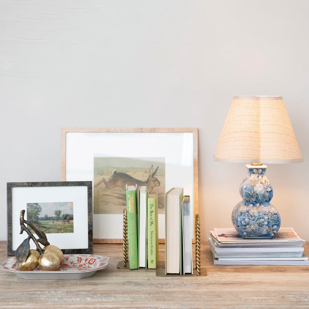 Books held by twisted brass bookends on a desk next to a blue patterned lamp and framed artwork.