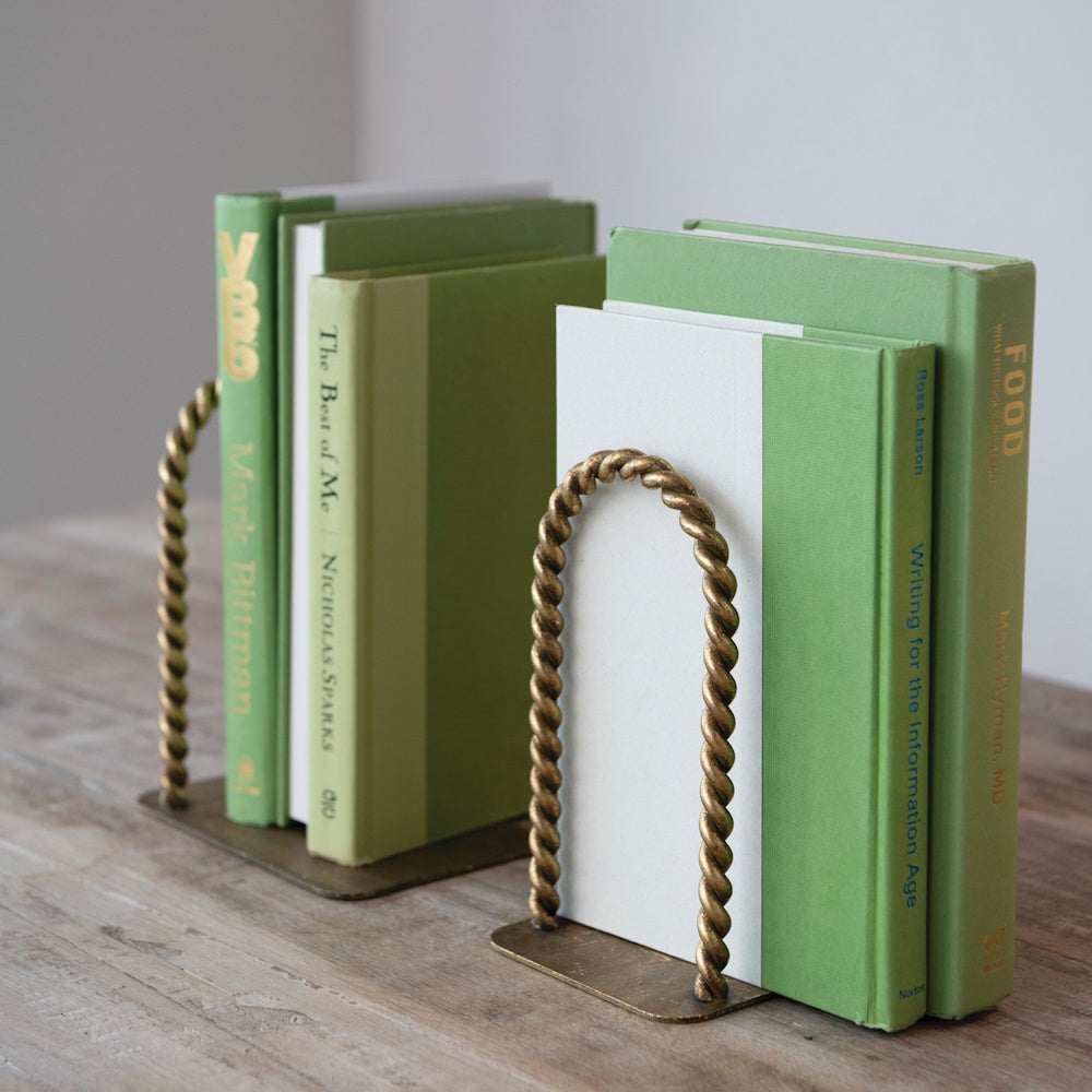 Two twisted brass arched bookends holding up rows of green-covered books on a rustic wooden table.