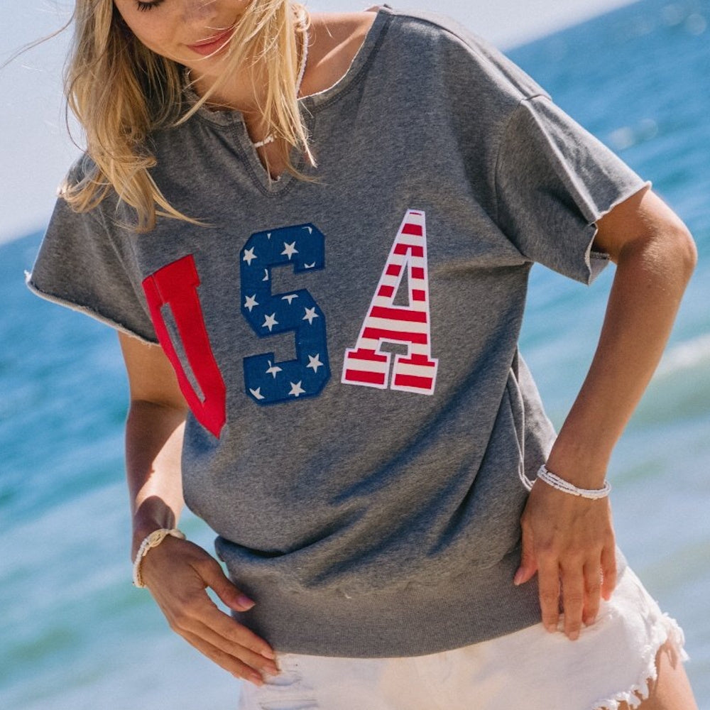 A woman at the beach wearing a grey short-sleeve top with "USA" in red, white, and blue embroidered letters.