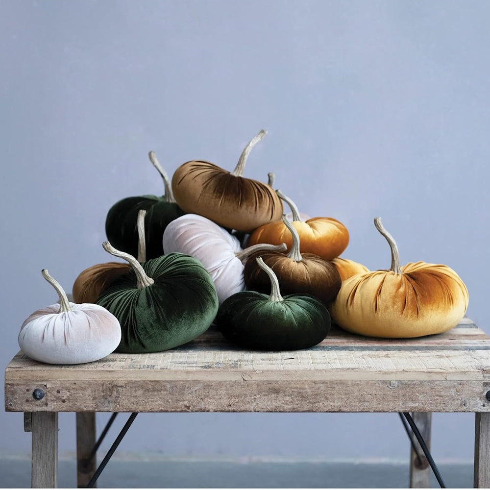 Plush velvet pumpkins in green, tan, and gold stacked on a rustic wooden table.