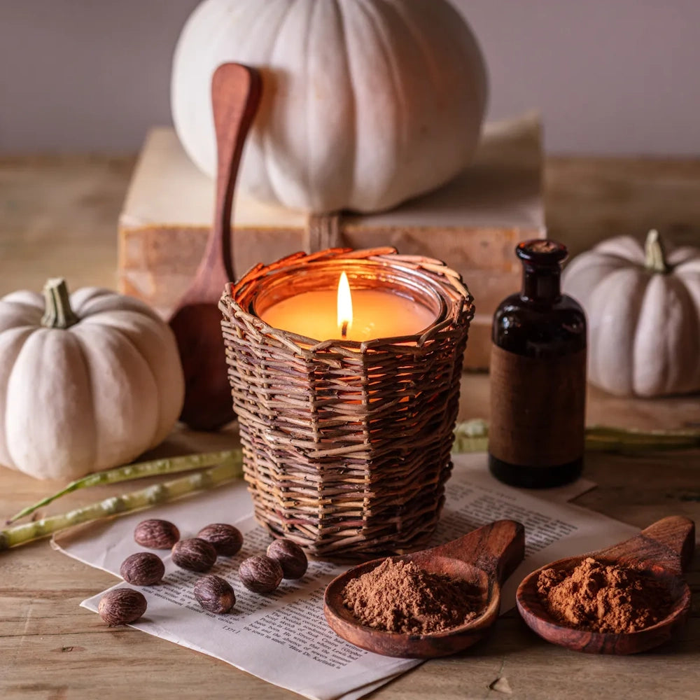 Lit candle in a woven basket holder on a rustic table with white pumpkins, spices, and a small glass bottle.