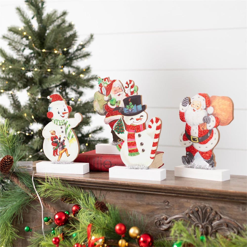 Vintage Santa and snowman wooden cutout table sitters on a mantel near a Christmas tree.