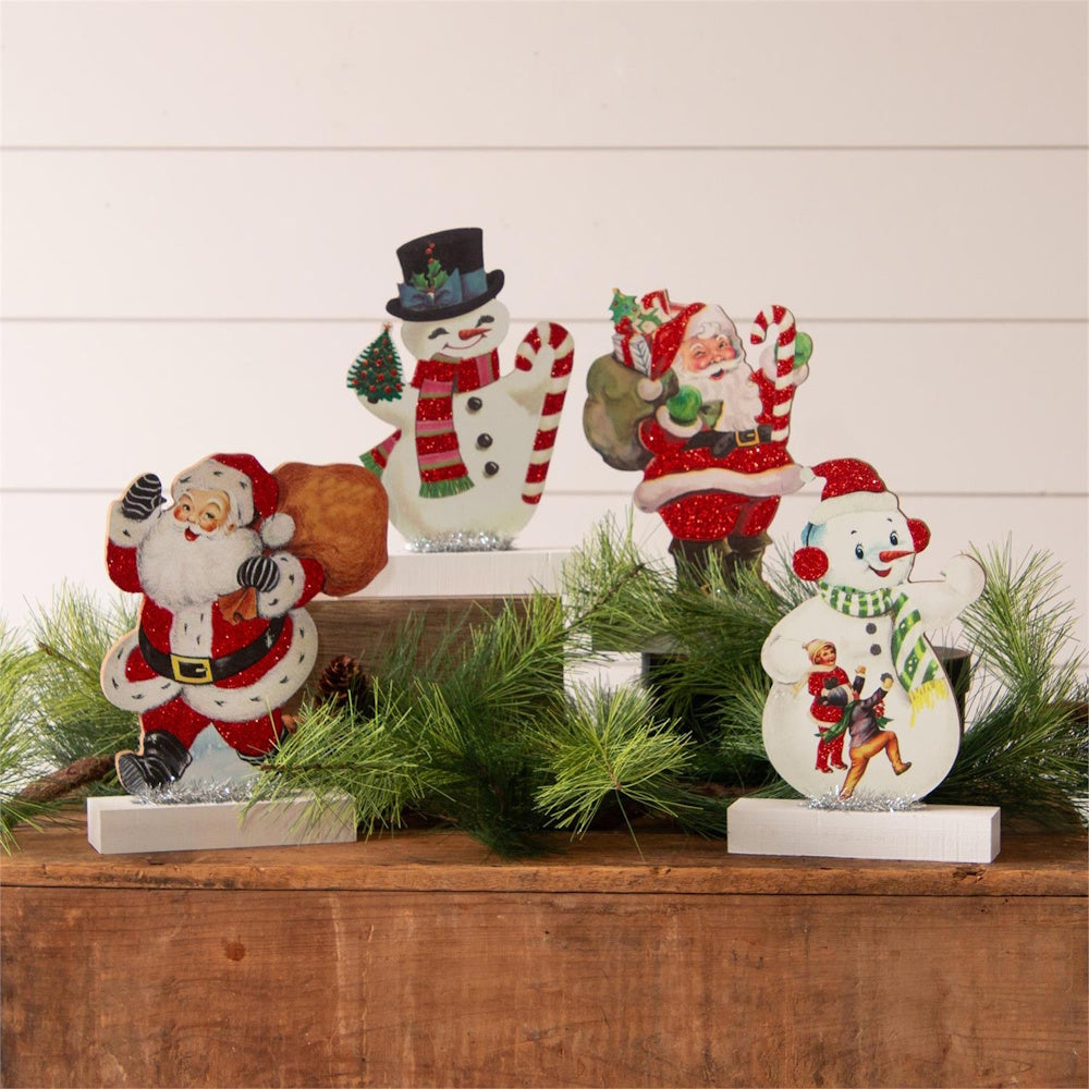 Four vintage-style Santa Claus and snowman cutout decorations displayed among green pine branches.
