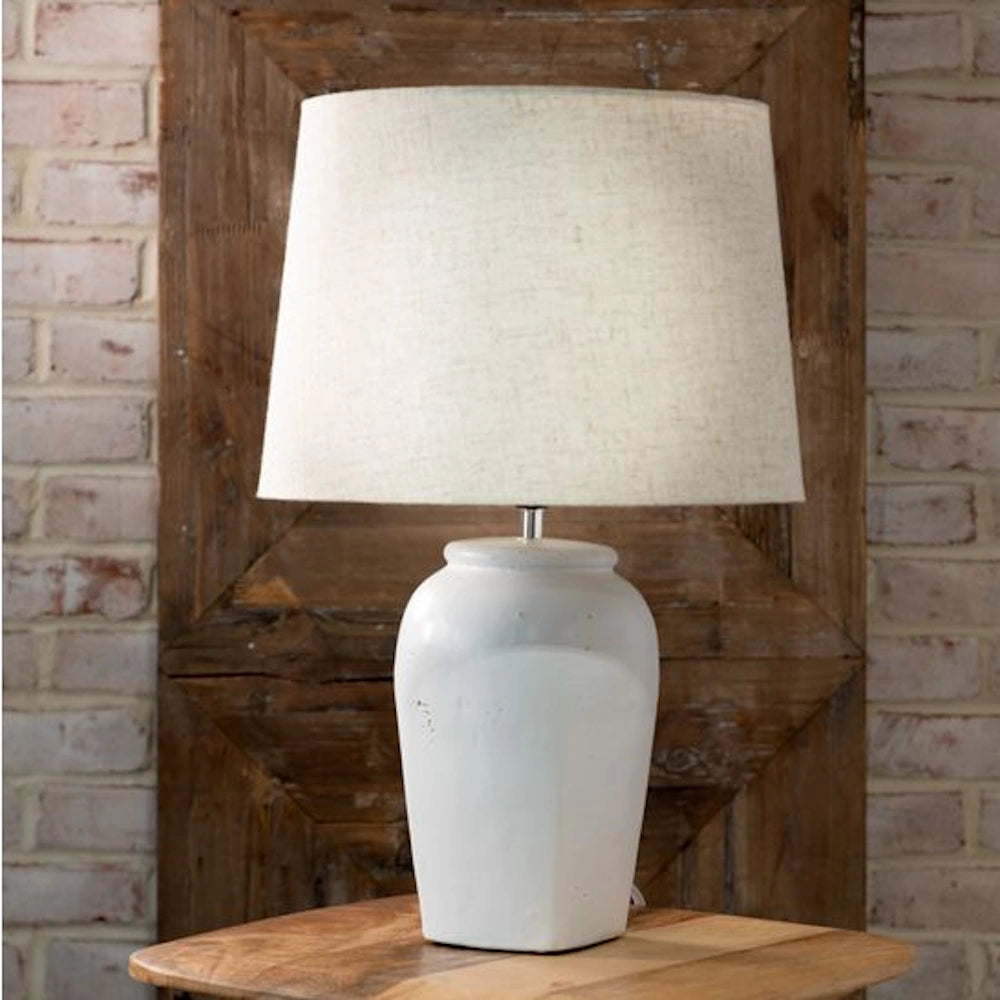 Vintage white ceramic jar table lamp with a beige linen tapered shade on a wooden table against a brick wall.