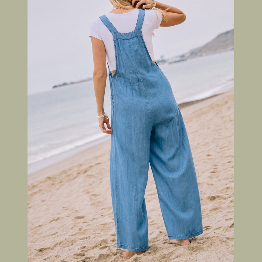 A woman from behind wears a light-blue chambray overall jumpsuit and white t-shirt on a sandy beach.