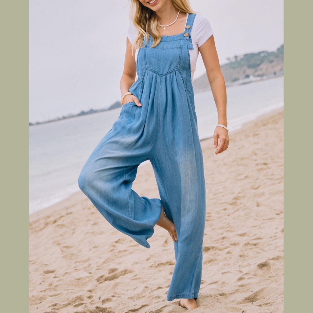 Woman posing on a beach wearing a light blue chambray overall jumpsuit over a white t-shirt.