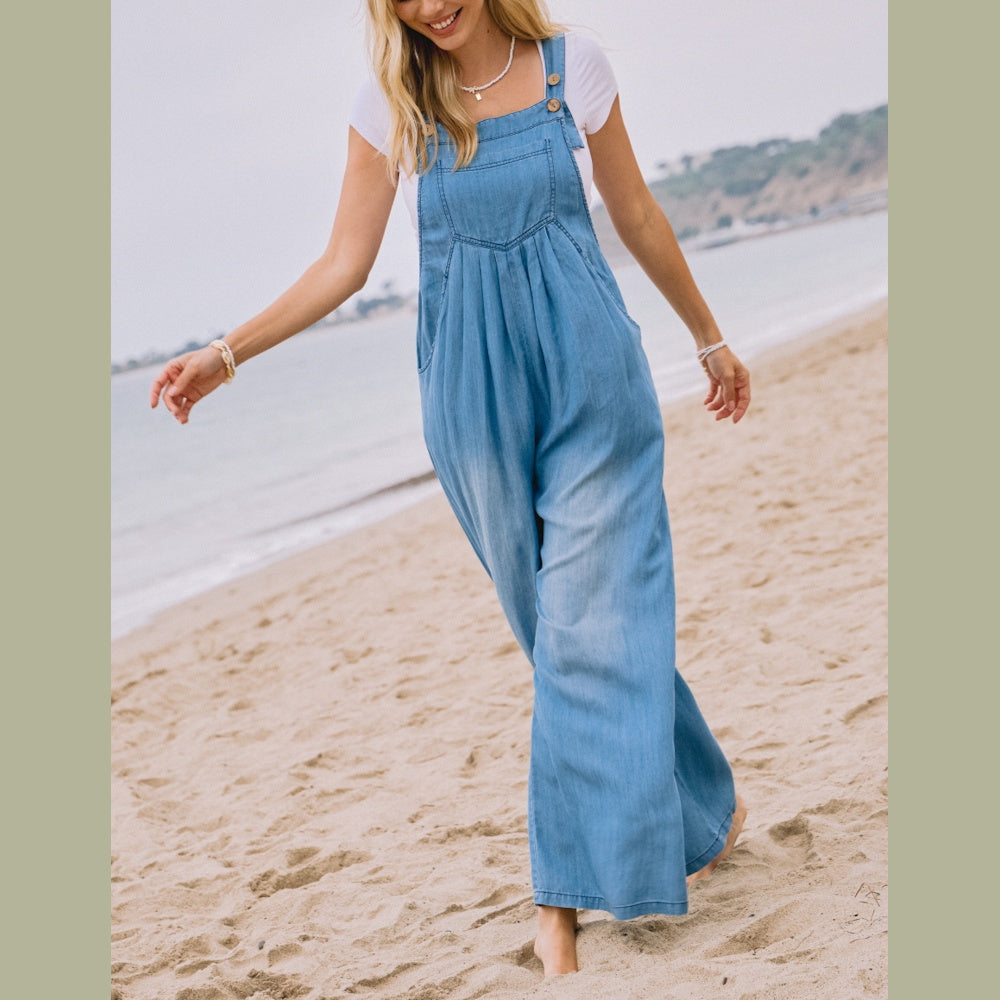 A woman walks barefoot on a beach wearing a light-blue chambray overall jumpsuit over a white T-shirt.