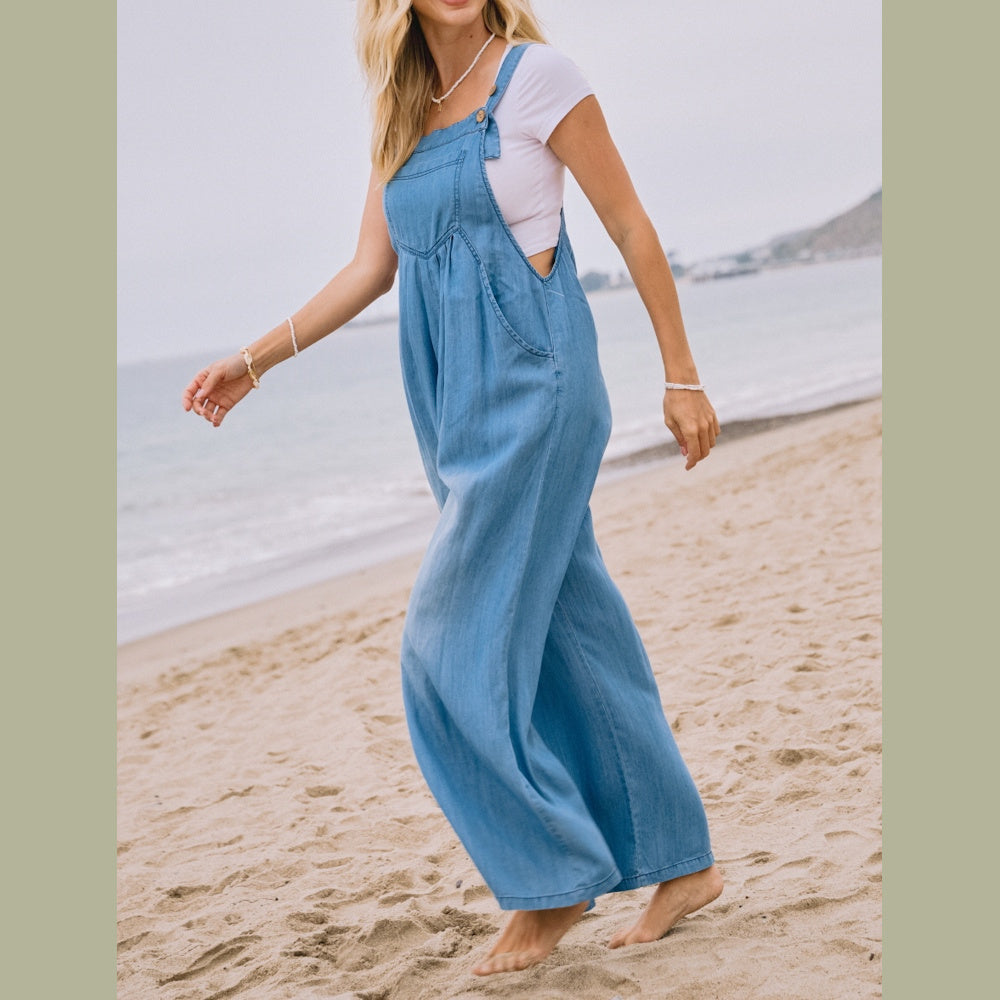 A woman walks barefoot on a sandy beach wearing a light blue chambray overall jumpsuit over a white t-shirt.