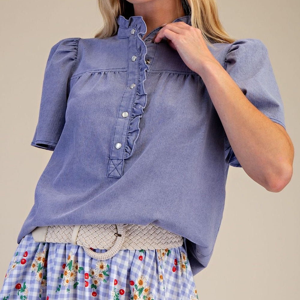 Front angle view of washed blue chambray short-sleeve top with ruffled trim and pearl snap buttons.