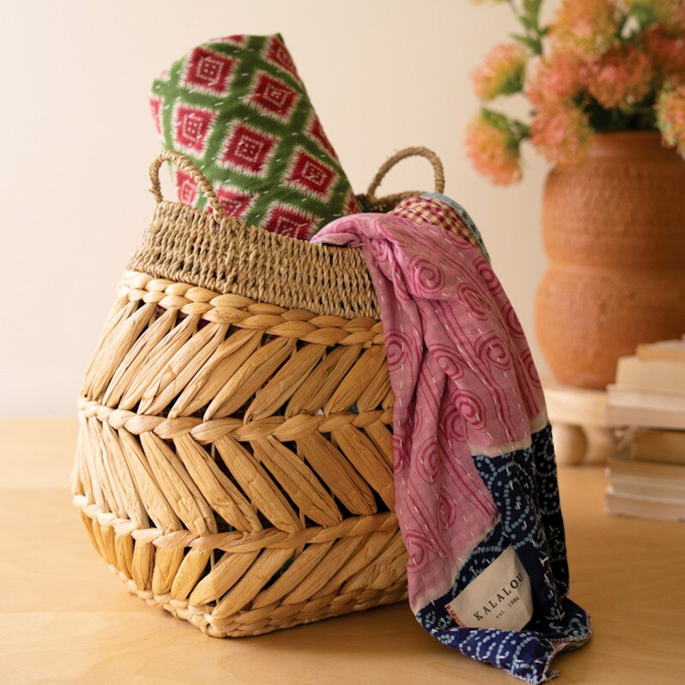 Woven water hyacinth basket holding colorful patterned textiles, with a pink scarf draped over the side.