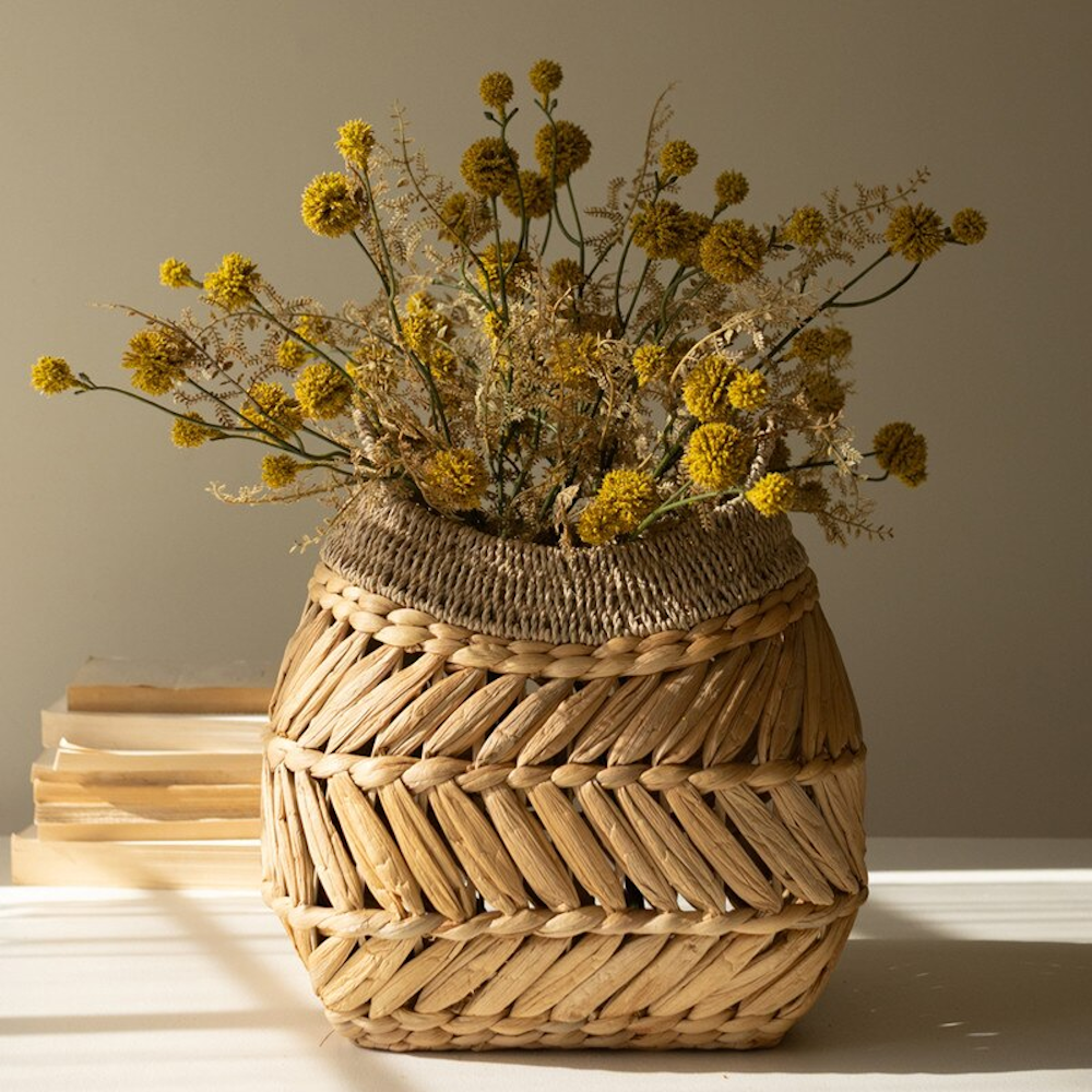 Woven water hyacinth basket vase filled with dried yellow Billy button flowers on a tabletop.