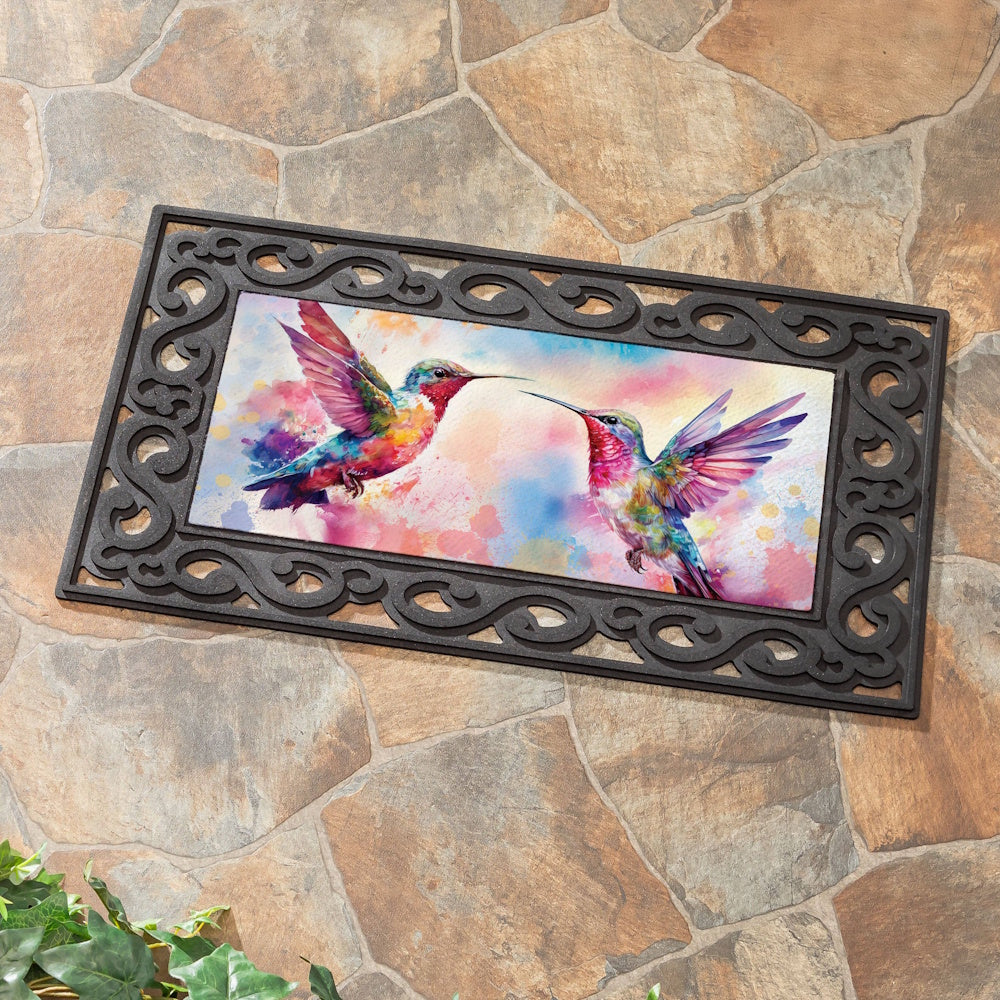 Watercolor hummingbirds on a decorative door mat with a black scrollwork border, set against a stone tile background.