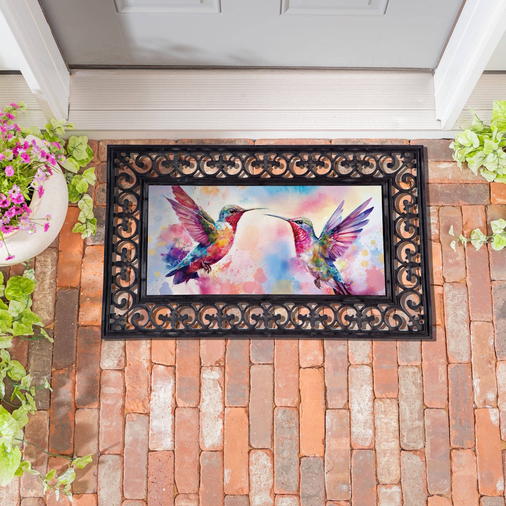 Watercolor hummingbirds on a decorative door mat insert, framed by a black scrollwork base on a red brick porch.