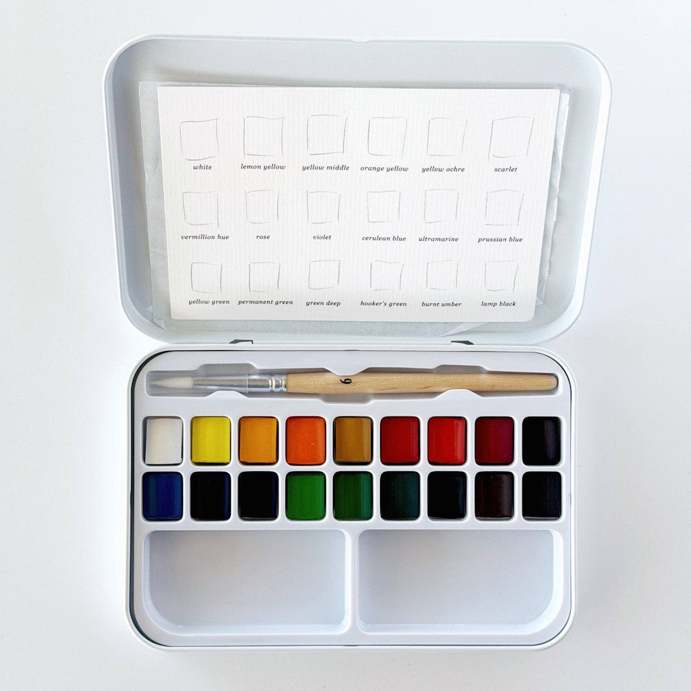 Open watercolor paint tin with a size 6 brush, 18 color pans, built-in mixing palette wells, and a swatch card inside the lid.