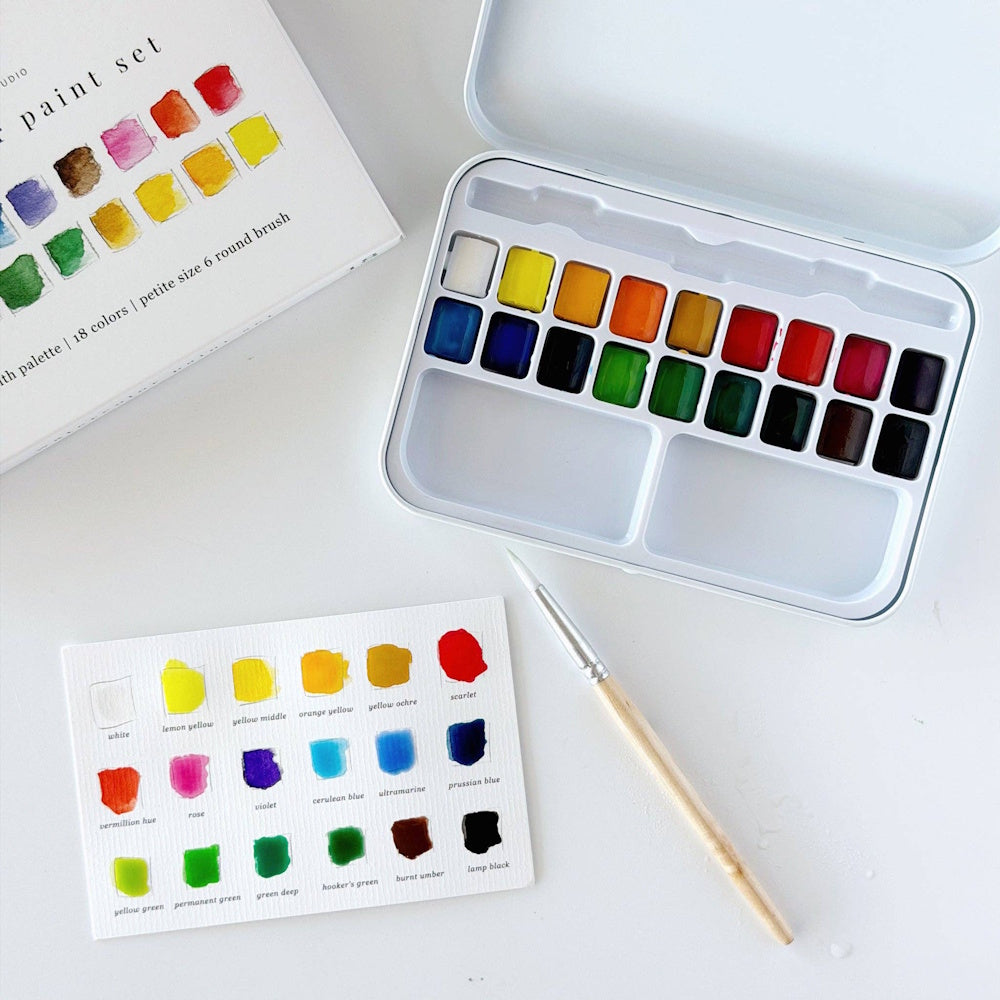 Open metal watercolor tin displayed alongside its outer cover box, a size 6 round brush, and a completed color swatch card filled in with paint.