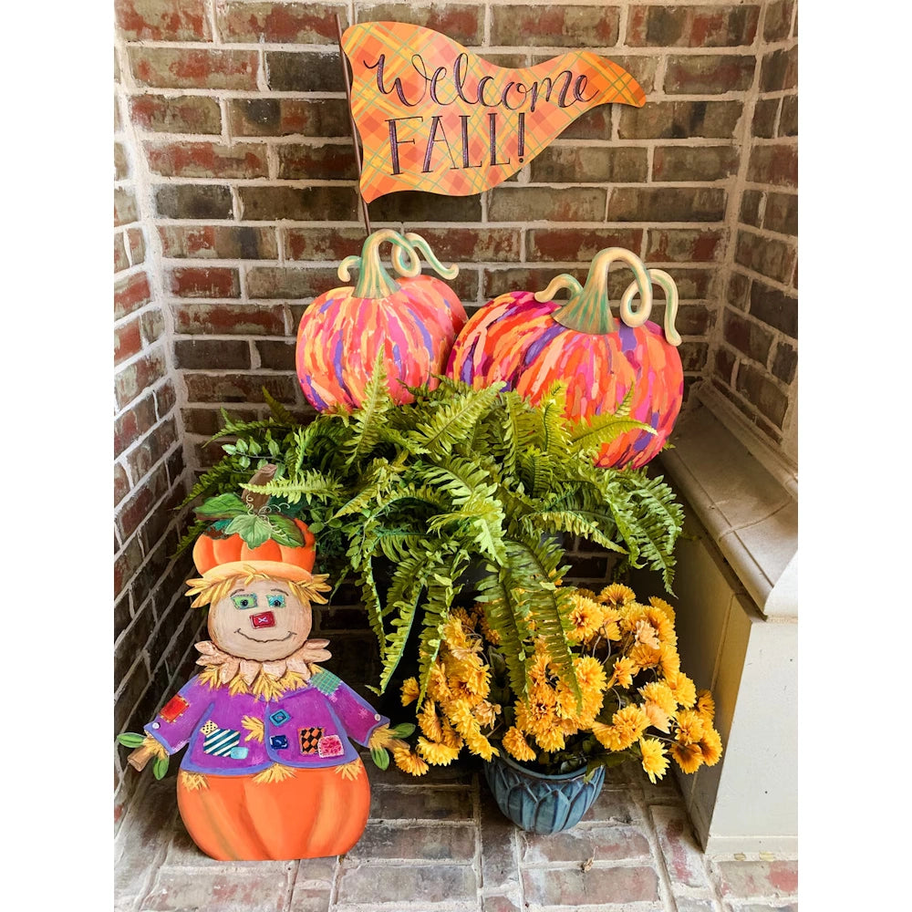 Autumn porch display with a scarecrow, colorful pumpkins, a Welcome Fall flag, yellow mums, and a fern.