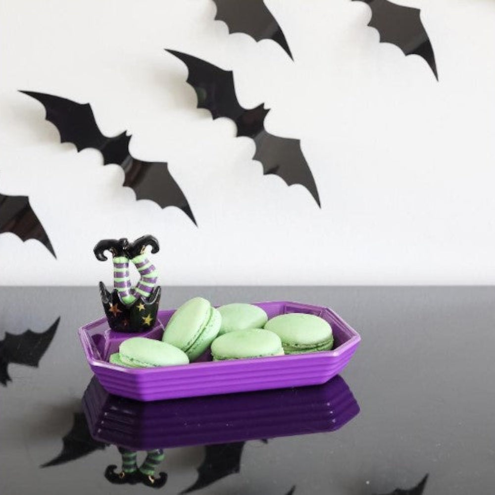 Green macarons in a purple Halloween tray with witch legs, below black bat cutouts on a white wall.
