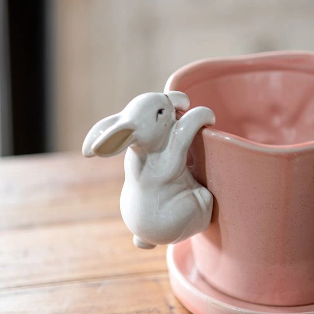 A white ceramic bunny pot hanger grips the rim of a pink glazed terracotta flower pot with a matching saucer.
