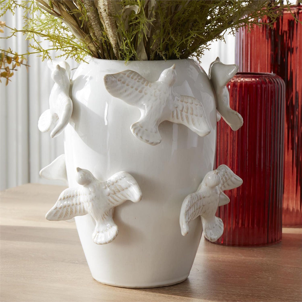 White ceramic vase with 3D flying bird accents and green foliage, styled next to red glass vases on a wood table.