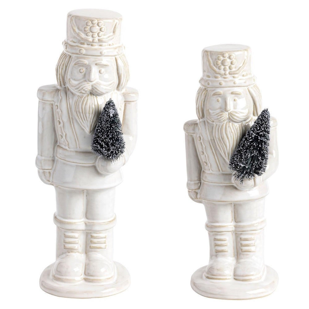 Two glazed white ceramic nutcracker figurines holding small, frosted bottle brush trees.