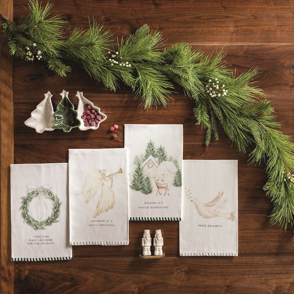 Four holiday tea towels displayed on a wooden surface with pine garland and festive decor.