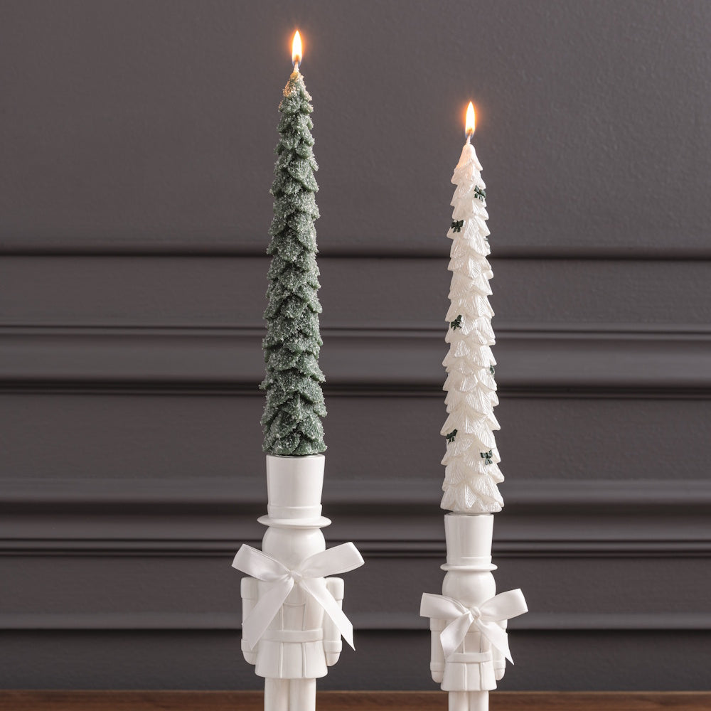 Lit green and white tree-shaped wax candles burning in white nutcracker holders, front view.