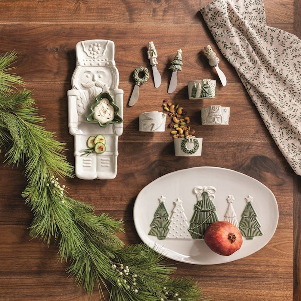 White ceramic Christmas dishes, nutcracker tray, tree platter, and knives on a wooden table.