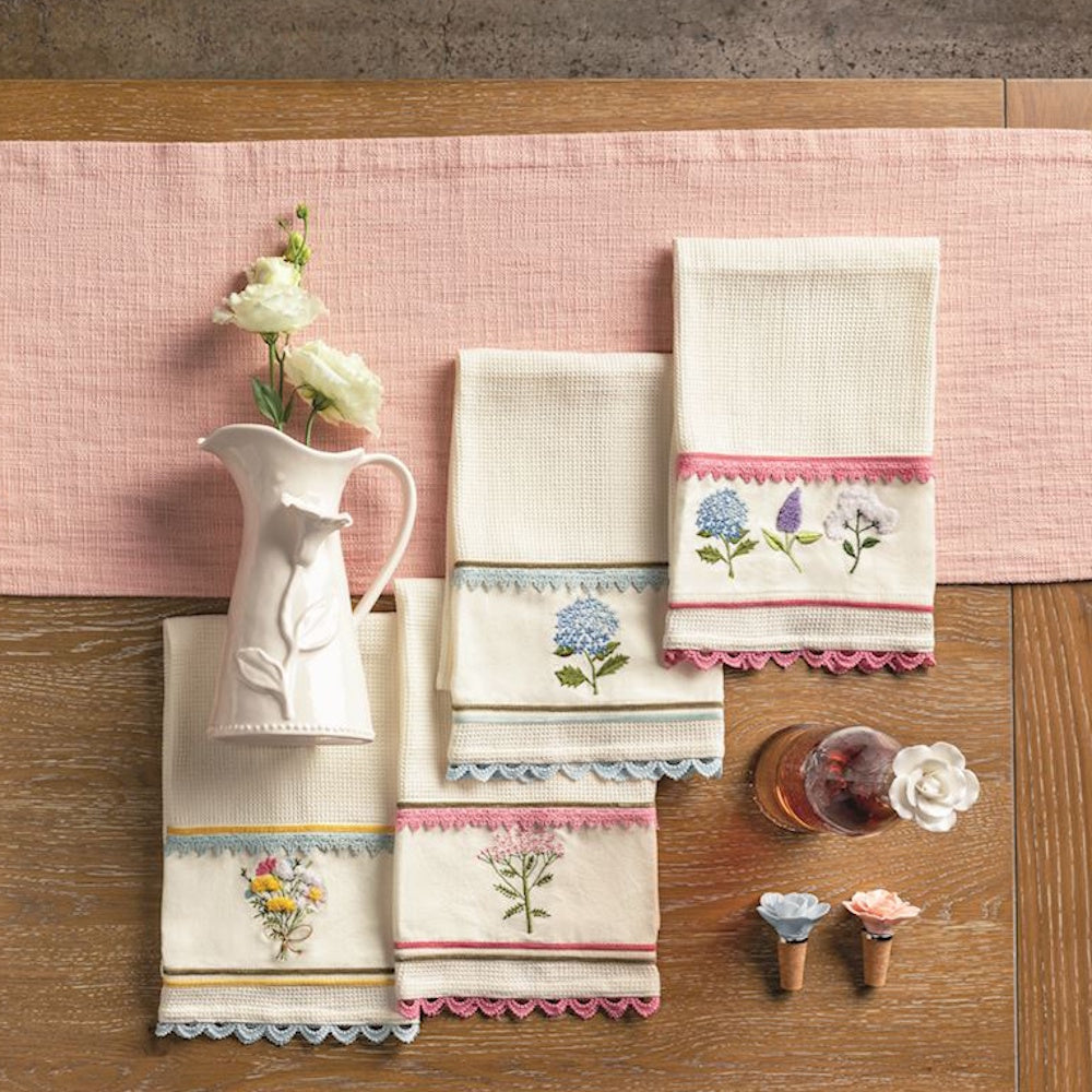 Embroidered floral tea towels and a white ceramic pitcher on a wooden table with a pink runner.