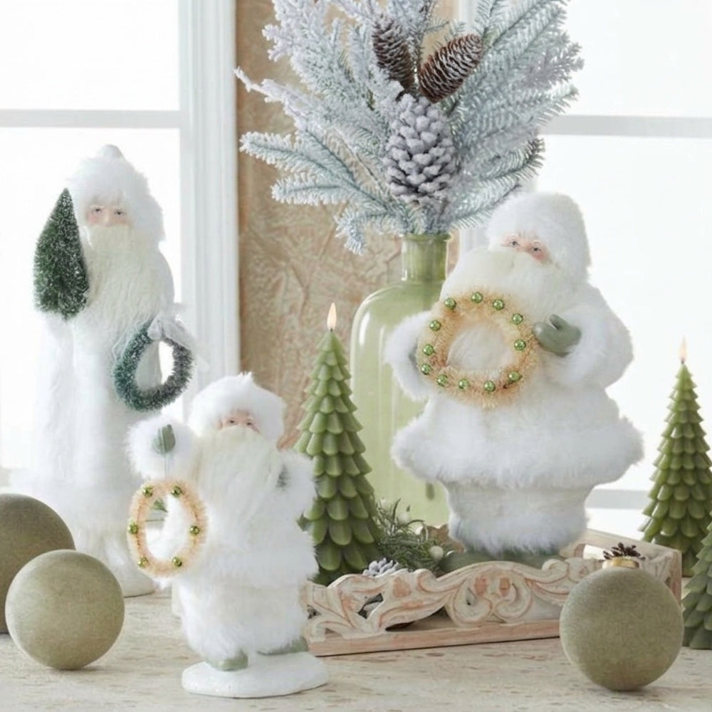 Lifestyle display of white faux fur Santa figurines holding wreaths, arranged with green tree candles, frosted pine stems, and decorative spheres.
