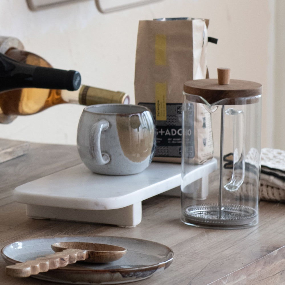 A white marble pedestal holding a ceramic mug and a bag of coffee next to a glass French press.