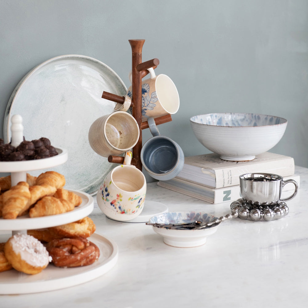 A white tiered tray filled with pastries next to a wooden mug tree holding ceramic mugs on a countertop.