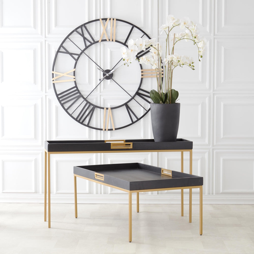 Black and gold tray tables displaying an orchid next to a large wall clock with Roman numerals.