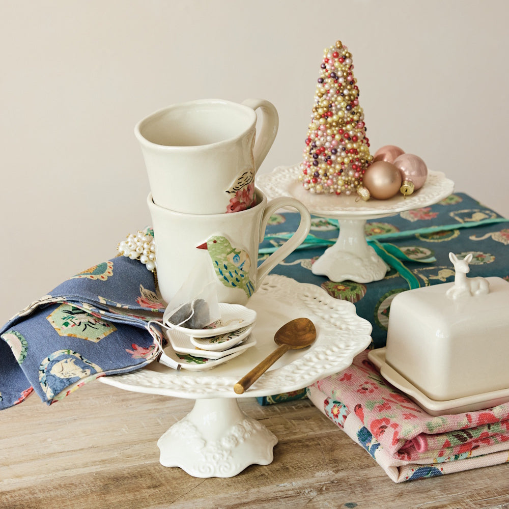 White stoneware pedestals holding stacked mugs and a beaded tree figurine on a decorated tabletop.