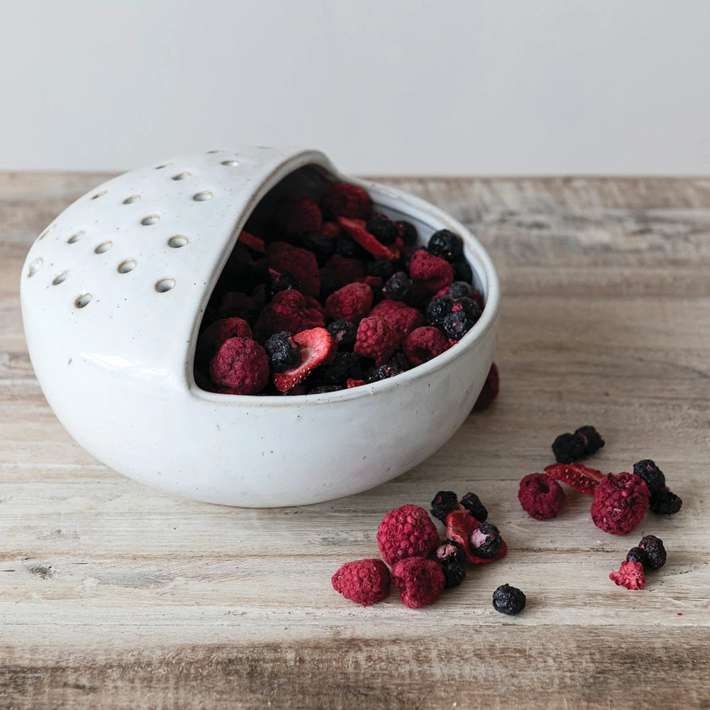 White stoneware strainer bowl filled with dried raspberries and blueberries on a rustic wooden surface.