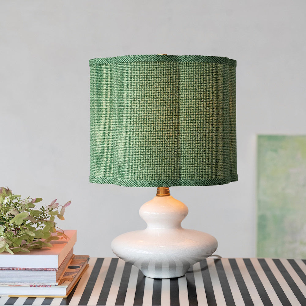 White stoneware table lamp with a scalloped green fabric shade on a striped surface next to books.