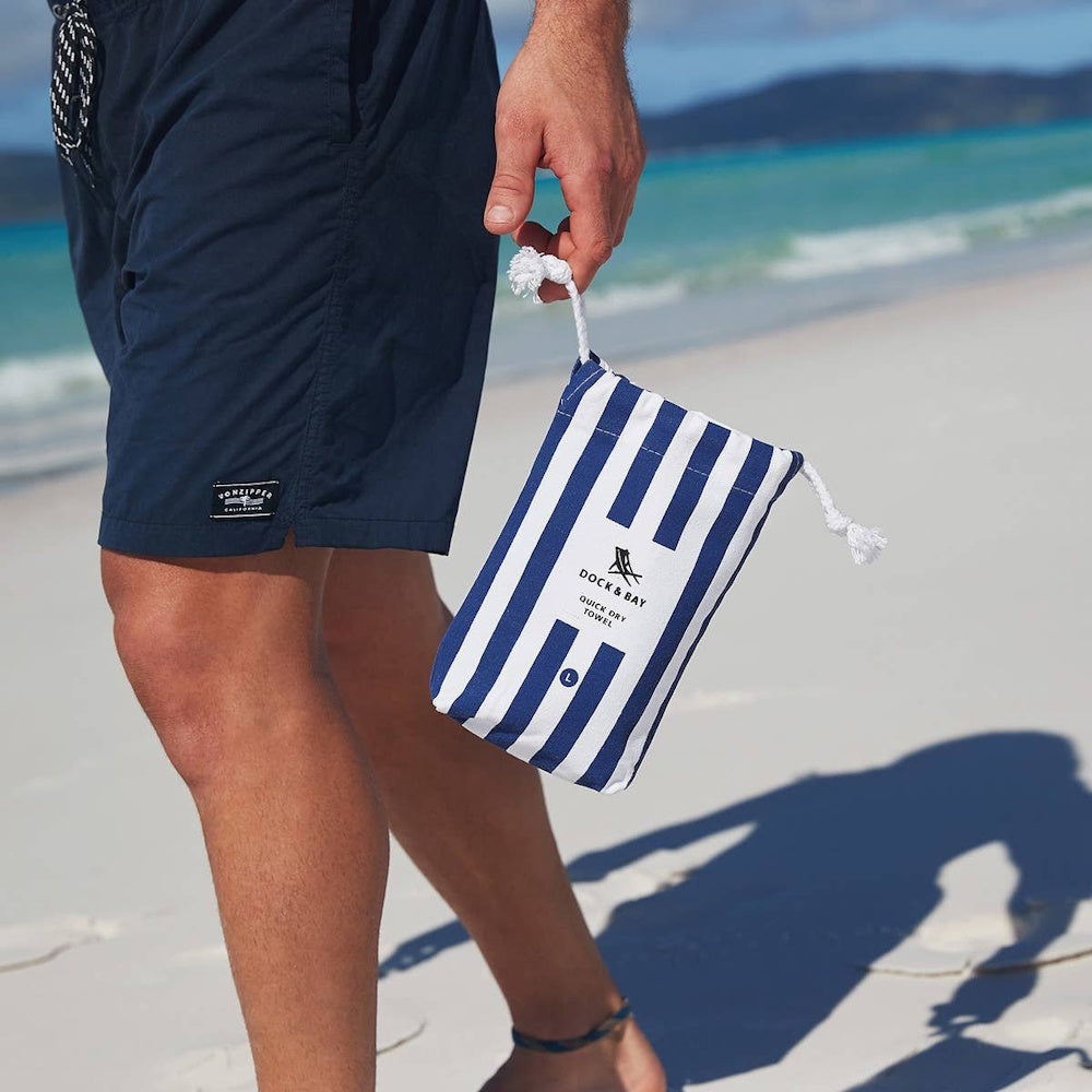 A person on a beach holding a blue and white striped Dock & Bay quick dry towel pouch.