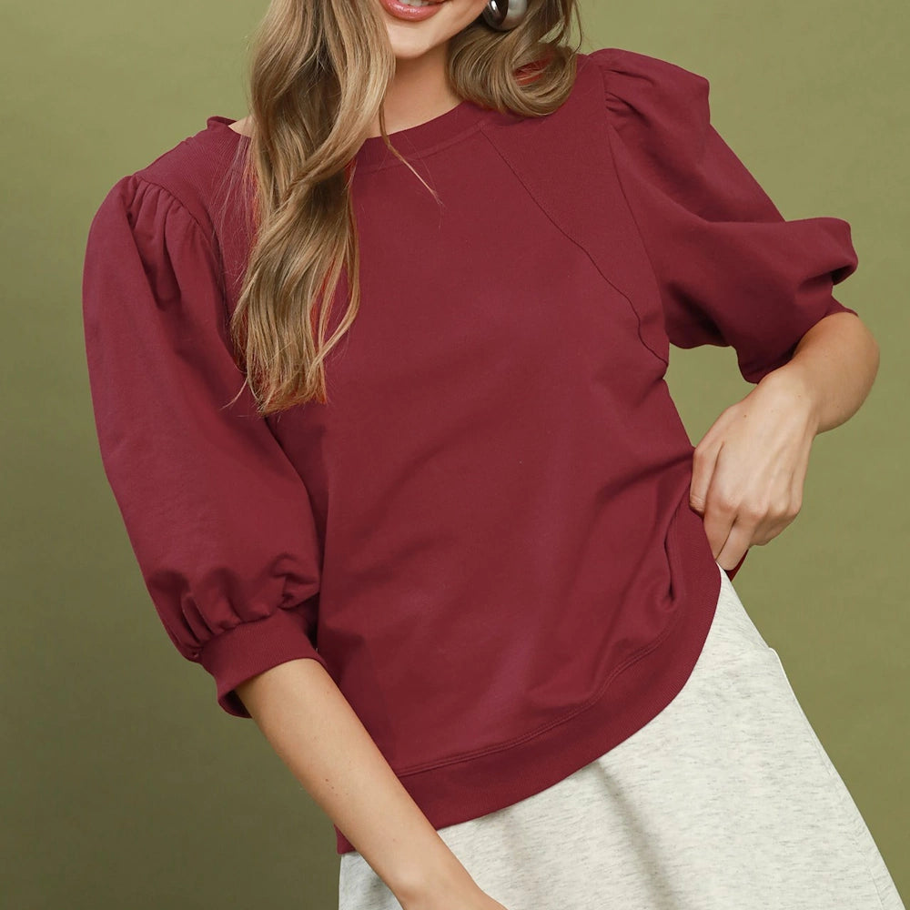 Woman wearing a wine-red crewneck sweatshirt with voluminous puff sleeves and ribbed cuffs.