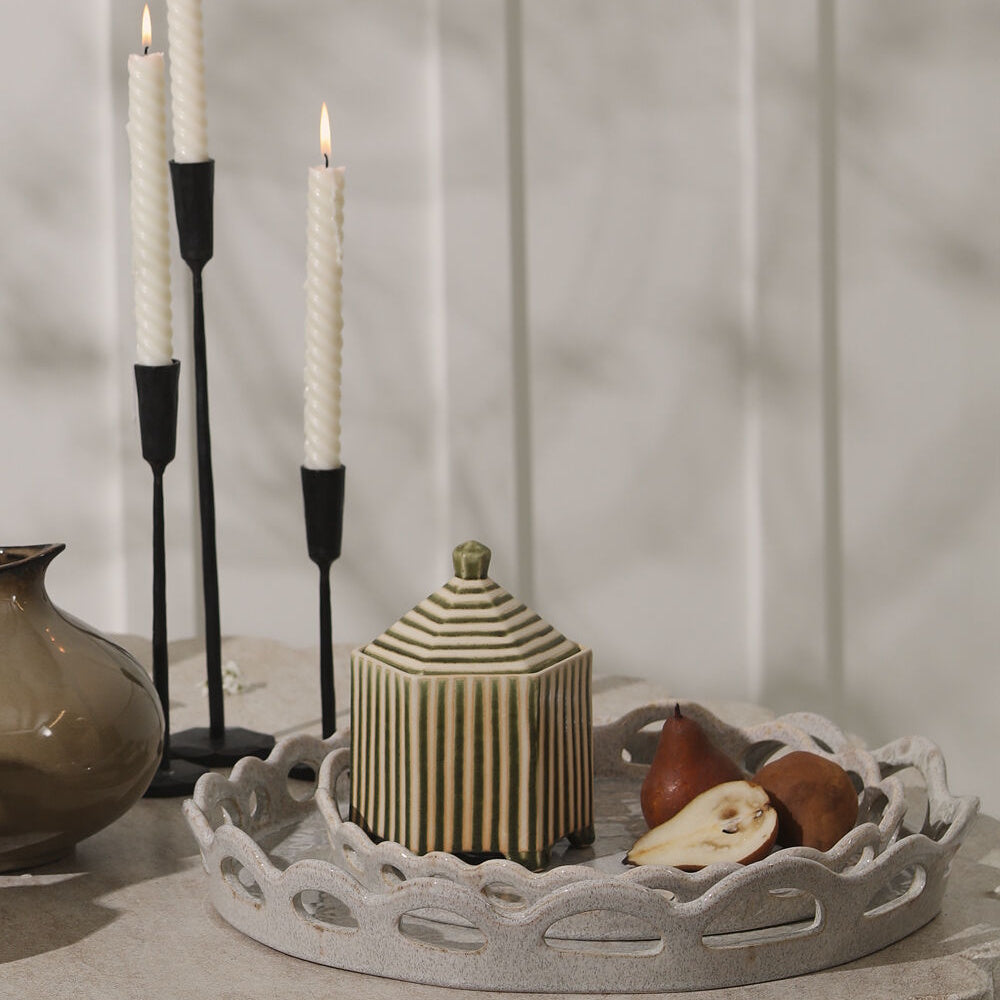 Ceramic trays holding a striped lidded jar and sliced pears, next to three burning taper candles.