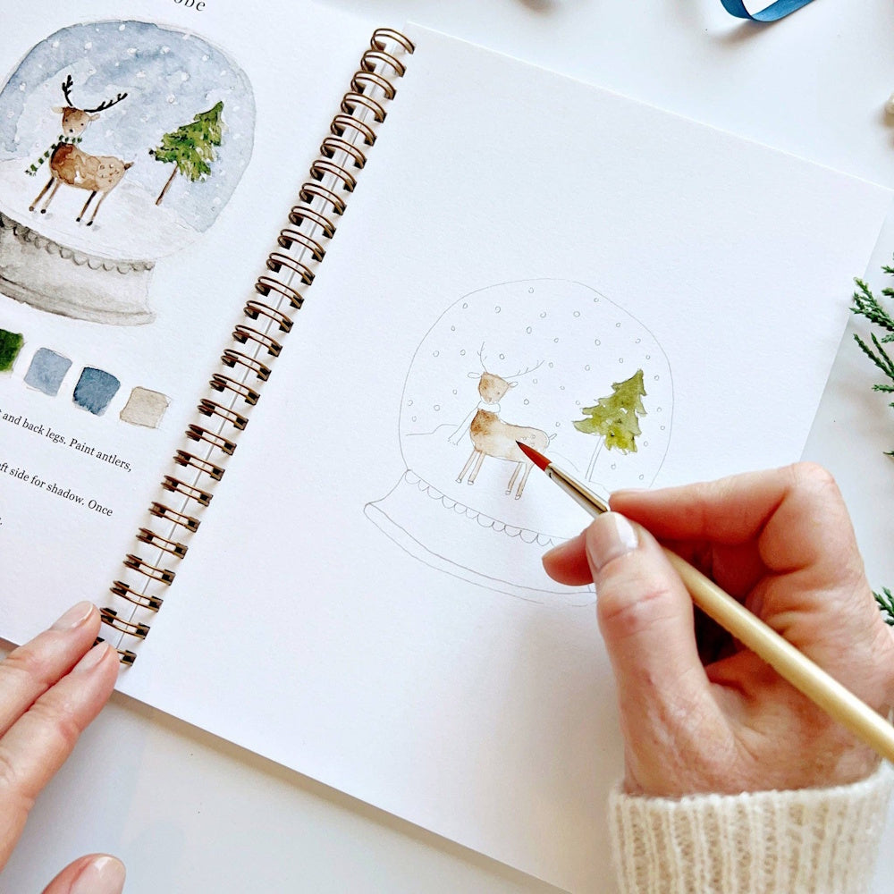 Hand using a paintbrush to fill in a snow globe deer illustration inside an open workbook.
