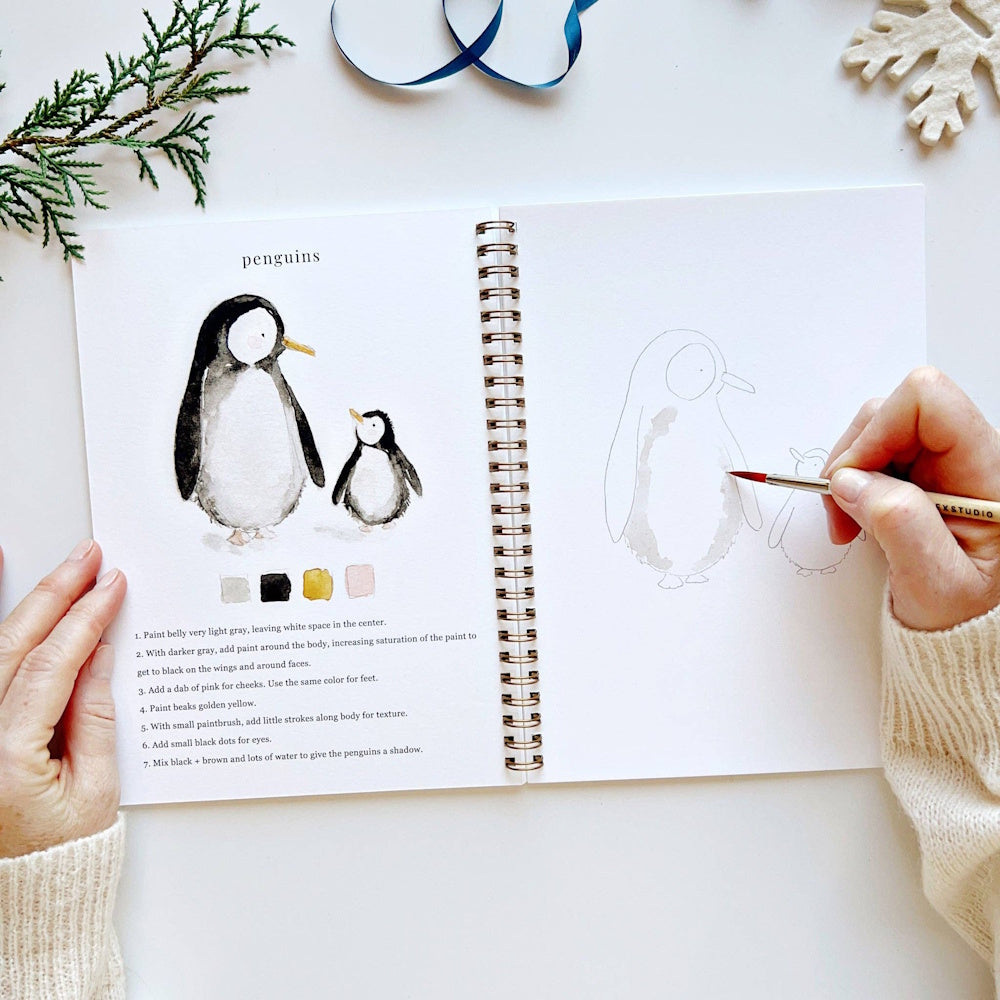 Open watercolor workbook showing step-by-step penguin instructions and someone painting the line art.