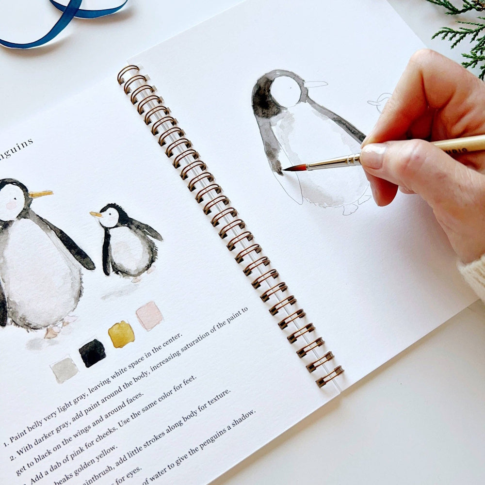 Hand using a paintbrush to add black watercolor paint to a penguin sketch in an open workbook.