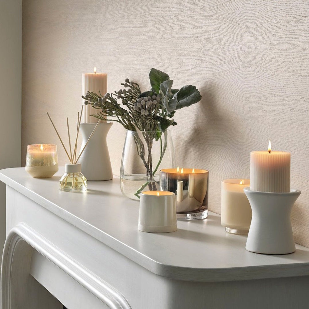 A mantle display featuring lit candles, pillar candles on stands, a reed diffuser, and a vase of greenery.