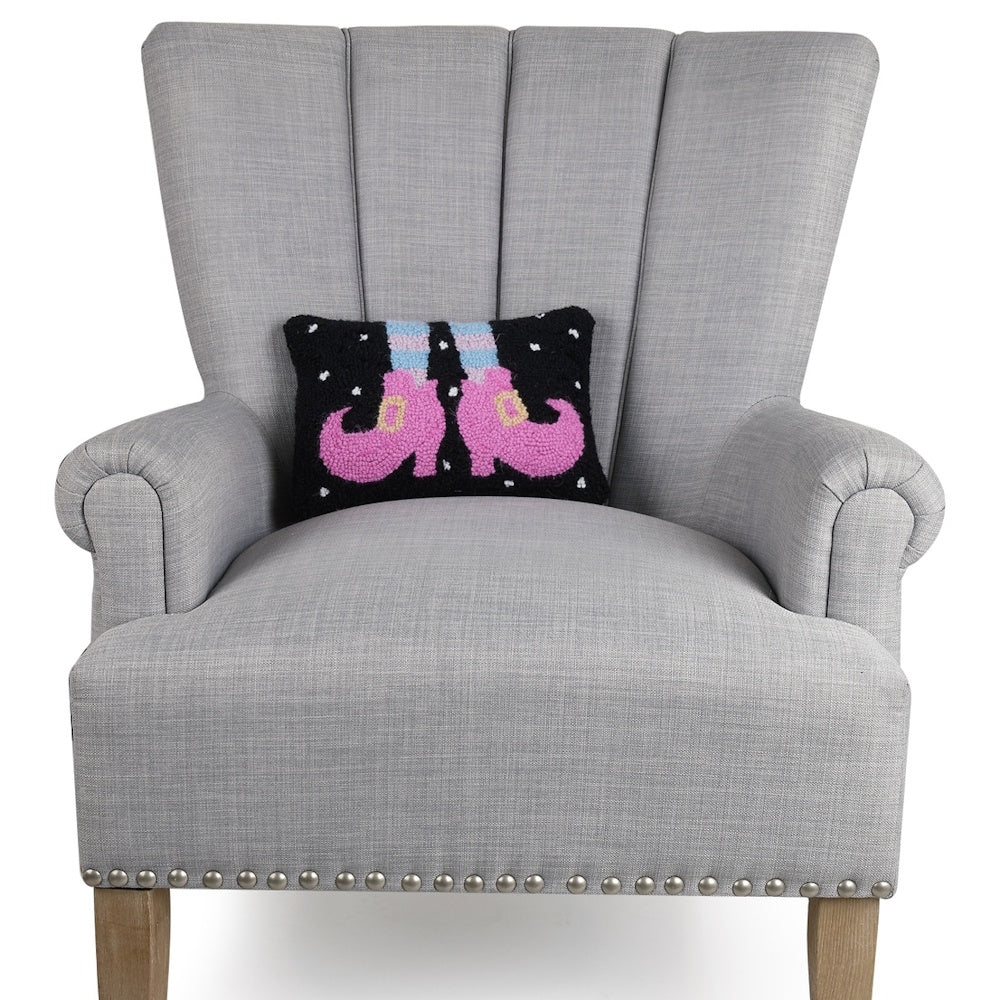 Black hooked throw pillow with pink witch shoes displayed on a gray armchair with nailhead trim.