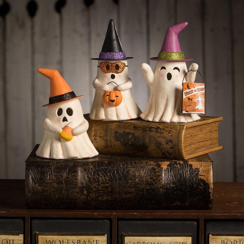 Three small ghost figurines wearing witch hats and carrying treats, displayed on old vintage books.