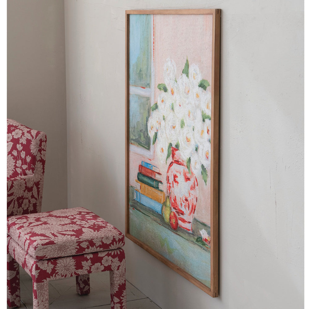 Floral painting in a wooden frame hanging on a wall next to a red patterned armchair and footstool.
