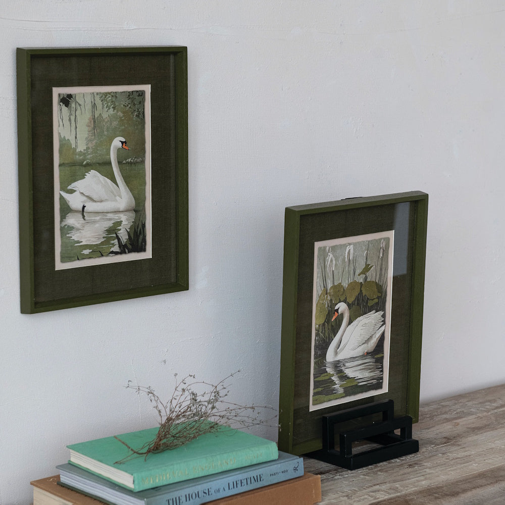 Two green framed swan prints displayed on a wall and tabletop next to books.