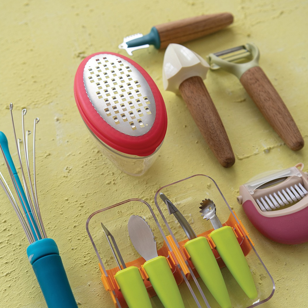 Assorted colorful kitchen tools including a grater, citrus reamer, whisk, and peelers on a yellow surface.