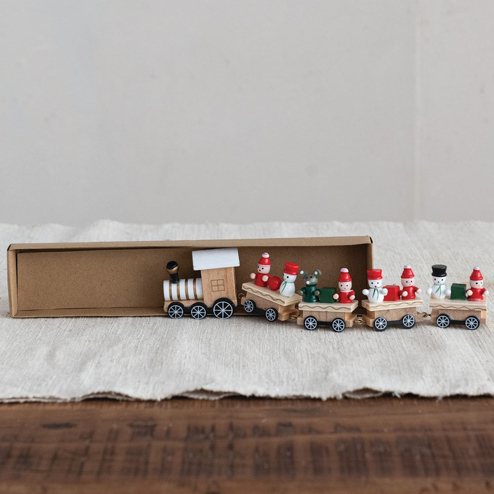 Miniature wooden toy train carrying festive holiday figures, pulling out of a brown cardboard box.